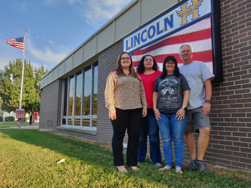 Four people in front of brick school with sign reading "Lincoln" and an American flag in the background