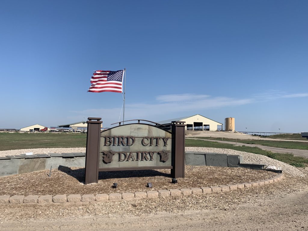Sign in the foreground reading "Bird City Dairy". Barn and American Flad against blue sky in the background.
