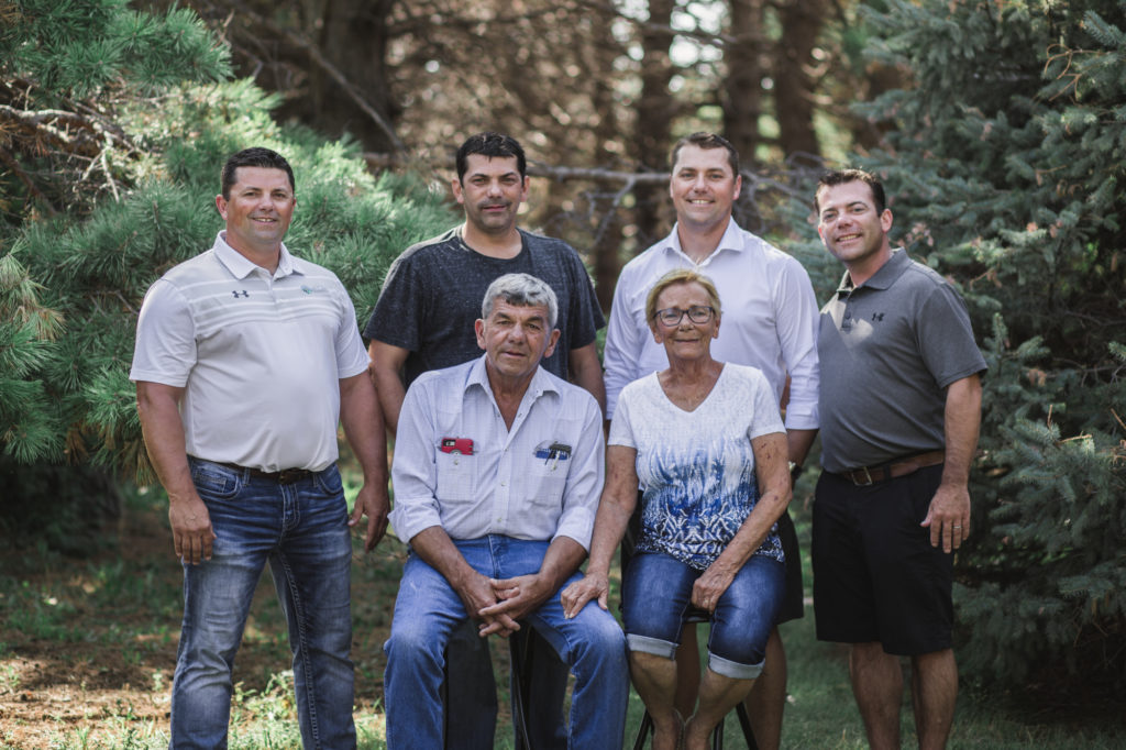 The McCarty Family in front of evergreen trees. Four adult sons in the back row, left to right: Mike, Clay, Ken, Dave. Front row, McCarty parents: Tom and Judy. 