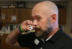 Image of Seth Dewey, Reno County Health Department's Substance Misuse Health Educator, holding Narcan nasal spray to his nose. He is demonstrating how Narcan nasal spray is administered to a person who has overdosed.