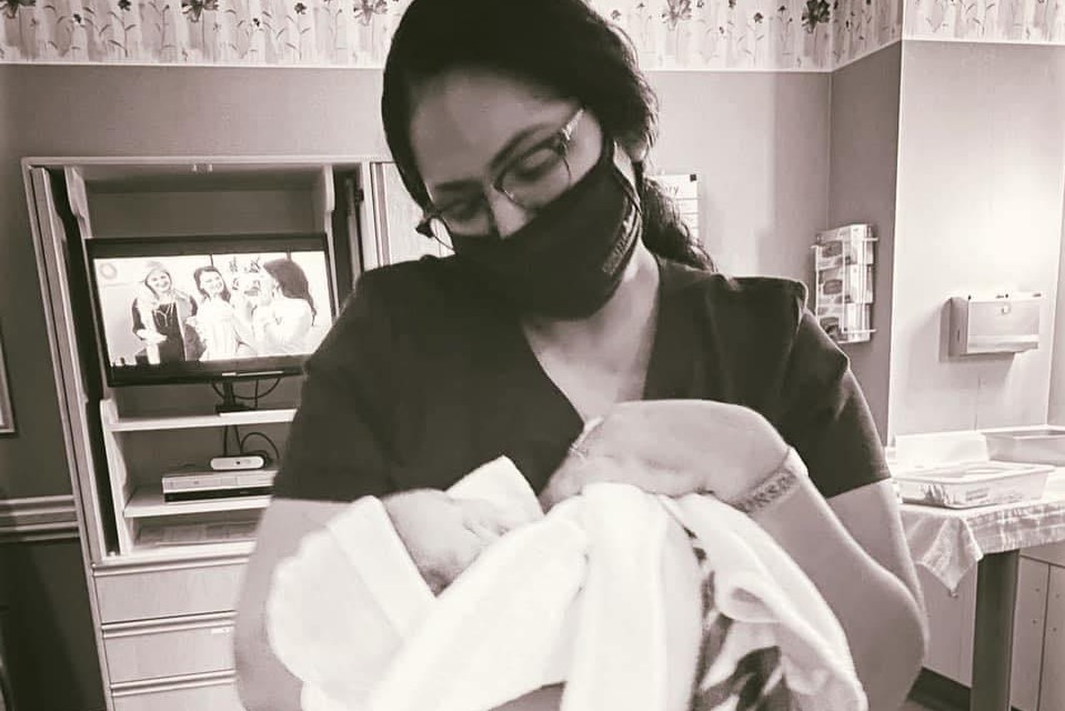 Image of a woman in a mask holding an infant swaddled in a blanket.